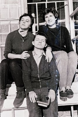 Three individuals pose casually on a concrete staircase in mid-20th-century attire. The man in the center wears a collared sh...