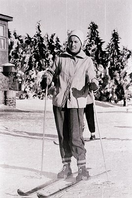 Mid-20th century skier in classic alpine attire—wide skis, long poles, and layered wool clothing—posing on groomed snow. Snow...