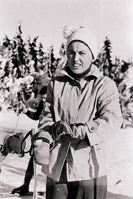 Mid-century alpine skier in vintage gear, standing on snowy slope with ski poles. White knit cap with pom-pom and insulated j...