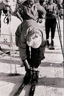 Mid-20th century skier in classic alpine stance, adjusting bindings on groomed snow. Heavy wool jacket, knit cap, and ski boo...