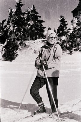 Mid-century cross-country skier in snowy forest, likely 1950s–1960s. Person wears classic ski attire: knit hat, goggles, padd...