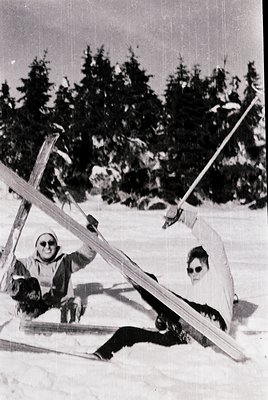 Two individuals pose playfully with vintage skis in a snowy forest, mid-20th century. Both wear sunglasses and winter attire;...