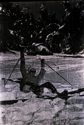 Vintage black-and-white ski jumper mid-air, arms outstretched on wooden skis, against a forested slope. Classic 1950s–1960s a...