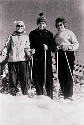 Three women pose on skis in mid-20th-century winter attire, likely 1960s–1970s. Snow-covered slope with wooden fence in backg...