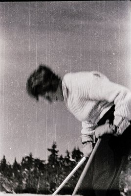 Mid-20th century black-and-white photo of a person skiing downhill, blurred motion capturing dynamic movement. Forest of ever...