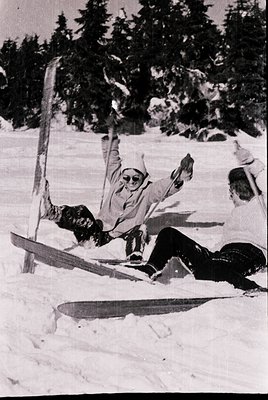 Black-and-white ski fall mid-1960s, alpine forest. Two skiers in vintage gear—wide skis, knee-high boots, and puffy jackets—l...