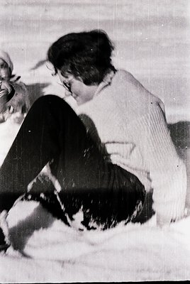Vintage black-and-white photo of a woman sitting on a snowy surface, petting a black-and-white dog. She wears a cardigan and ...