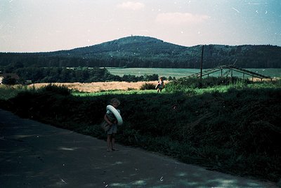 Vintage black-and-white photo of rural countryside scene with two children near a roadside. One child holds a white bag, whil...