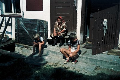 Vintage 1970s street scene featuring three individuals in a courtyard. Adult in patterned shirt and red headscarf sits on ste...
