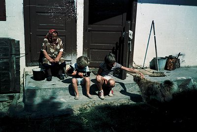 Vintage rural scene featuring an elderly woman in traditional attire seated on a concrete step, interacting with two children...