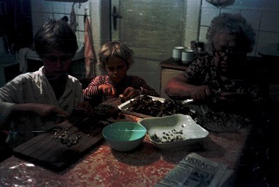 Three individuals in a dimly lit kitchen, likely mid-20th century, preparing food. The central figure, a child, assists with ...