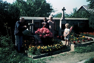 Family tending to a grave in a rural cemetery, 1960s-1970s. Man in dark jacket waters vibrant flowers; woman in floral dress ...