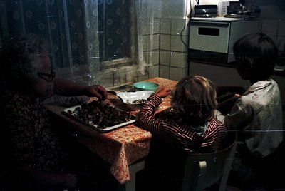 Vintage kitchen scene featuring three generations: an elderly woman (left) preparing food on a tray, a child (center) seated ...