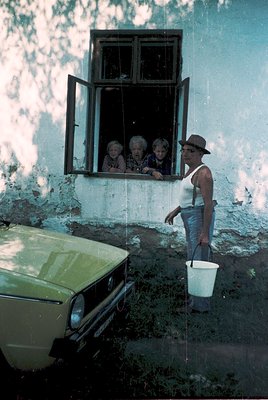 A man in a fedora and sleeveless shirt holds a white bucket, washing a weathered wall beside a vintage green car. Three elder...