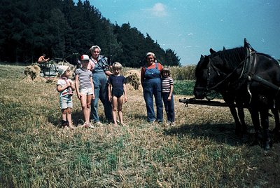 Family poses beside a horse-drawn hay wagon in a rural field, 1960s-1970s. Adults in overalls and striped shirts, children in...