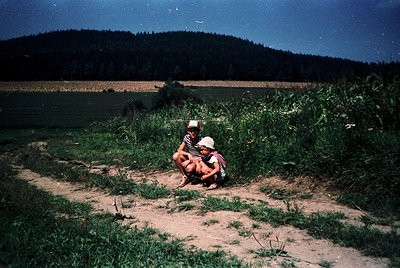 Two individuals sit on a dirt path surrounded by wild grass and crops under a starry night sky. The person on the left wears ...