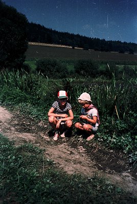 Two children sit on a dirt path in a rural countryside setting, surrounded by tall grass and trees. The girl on the left wear...