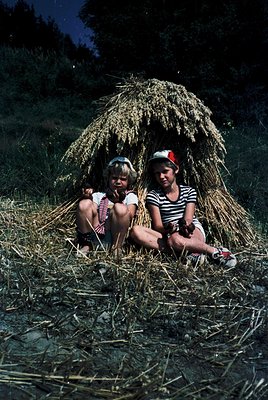 Two children pose under a haystack in a rural field, likely mid-20th century. The girl on the left wears a striped dress with...