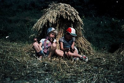 Two children sit beneath a thatched hay shelter in a rural field, mid-20th century. Boy wears red-and-white striped shirt, ca...