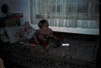 Child sits on patterned carpet in dimly lit room, holding a toy gun. Surrounding objects include a striped pillow, a plastic ...
