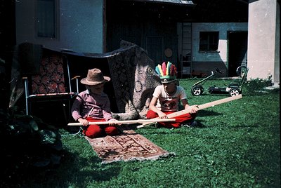 Two individuals pose outdoors in traditional Indigenous attire, seated on woven mats. The adult wears a wide-brimmed hat and ...