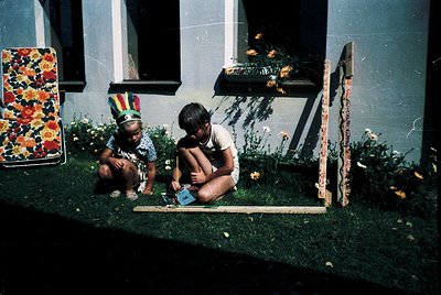Two children play with a wooden board game on grass beside a house, 1970s. One wears a floral-patterned headband; the other s...