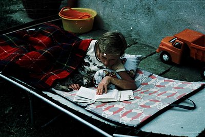 Child reading outdoors on a vintage metal camping table, surrounded by mid-20th-century camping gear. Red and black striped s...