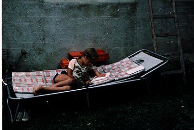 Child sits on a metal camp cot in an outdoor or semi-enclosed space, holding a small object (likely a toy or tool). The cot h...