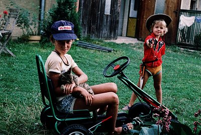 Vintage childhood scene: two children in 1970s-style outdoor play gear—blue cap with "Vodol" logo, floral shirt, and a girl i...