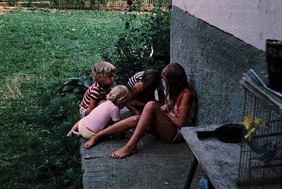 Four children sit on a concrete ledge in a grassy, shaded area, engaged in conversation. The girl on the right wears a red he...