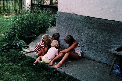 Three children huddle on a concrete step in a lush garden, mid-1970s. Left wears striped shirt, center in pink dress, right i...