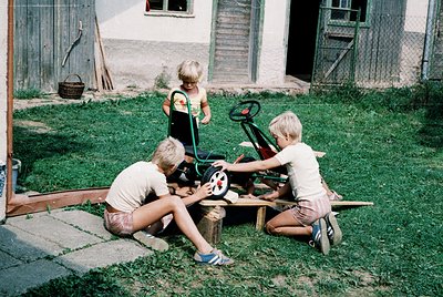 Three children play with a toy tractor in a backyard, c.1970s. The boy on the right wears a white T-shirt and shorts, kneelin...