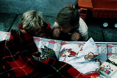 Two children sit closely on patterned bedding, engrossed in a shared activity. The girl wears a red sweater, the boy a white ...