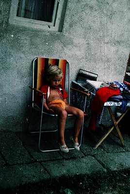 Child in vintage deck chair against weathered concrete wall, mid-20th century urban setting. Patterned striped fabric and ora...