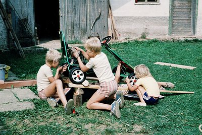 Three boys in 1970s-style shorts and tank tops repair a vintage green lawnmower on grass near a wooden barn. Mid-century rura...