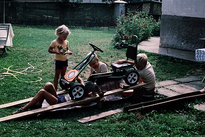 Vintage backyard scene: two children and an adult assembling a homemade lawnmower cart on wooden planks, 1960s-70s. Bright su...
