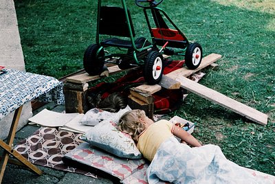 Vintage toy lawnmower and child resting on patterned blankets in grassy outdoor setting. Mid-20th century play scene evokes n...