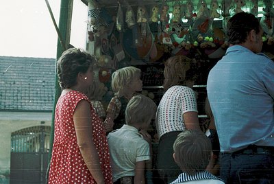 Family group examines colorful ceramic souvenirs at a seaside stall, likely mid-20th century. Bright polka-dot dresses and st...