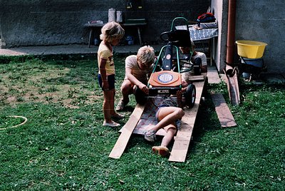 Vintage backyard scene featuring three children (1960s-1970s) with a homemade go-kart on wooden planks. Child in foreground r...