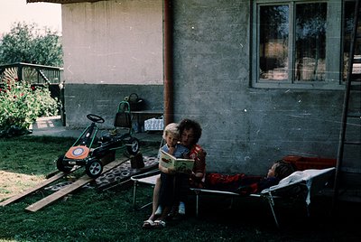 Vintage mid-century backyard scene with two children: one seated on a metal bench reading a book, another reclining on a loun...