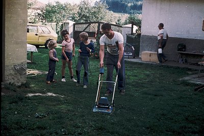 Family mows lawn in suburban backyard, 1970s. Man pushes reel mower; children stand nearby in casual summer attire. Yellow ca...