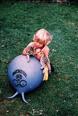 Vintage-style photo of a young child (mid-1970s) hugging a striped kitten atop a blue "Montezuma" brand exercise ball in an o...