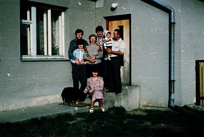 Family portrait outside a Soviet-era apartment building, likely Eastern Europe, 1970s–1980s. Six individuals pose on concrete...