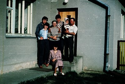 Family portrait outside a concrete residential building, likely Eastern Bloc-era (1970s–1980s). Five adults and one child pos...