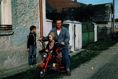 Vintage 1970s rural scene: man in striped sweater assists child on red pedal cart, another child stands nearby. Weathered pla...