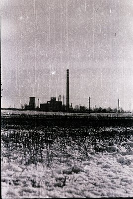 Mid-20th century industrial landscape featuring a smokestack and factory complex. Snow blankets the foreground, likely indica...