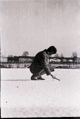 Mid-20th century black-and-white photo of a man shoveling snow in an urban setting, likely . He wears a long coat, trousers, ...