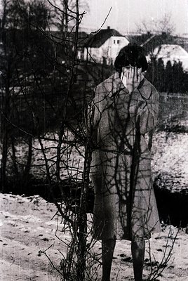 Black-and-white portrait of a child in winter attire, standing barefoot in snow. The child’s reflection is mirrored in leafle...