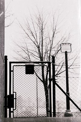 Vintage-style black-and-white photo of a gated entrance featuring a chain-link fence, metal gate, and vintage street lamp. Th...