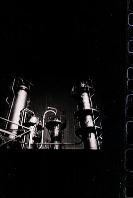 Industrial machinery with vertical pipes, valves, and metal frameworks in mid-20th century monochrome. Likely a factory or re...